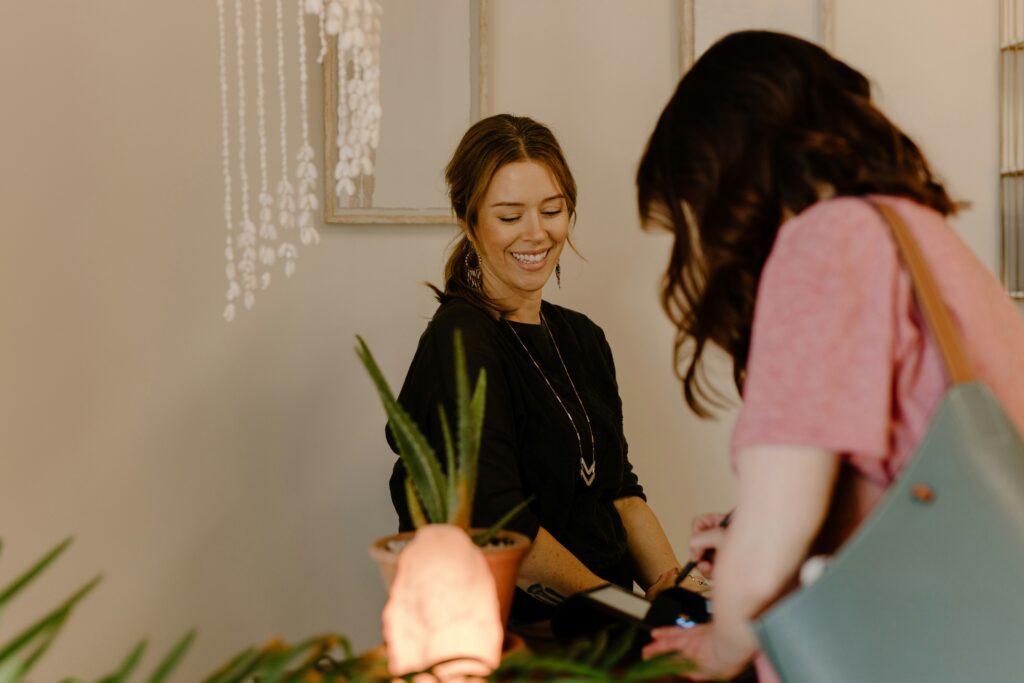 A warm lifestyle shot of a smiling salon receptionist with brown hair tied back, wearing a black top and a long necklace. She is looking down as a client in a pink shirt completes a transaction on a card machine at a wooden counter. The scene is decorated with a potted aloe vera plant and a glowing Himalayan salt lamp, creating a welcoming and calm environment.