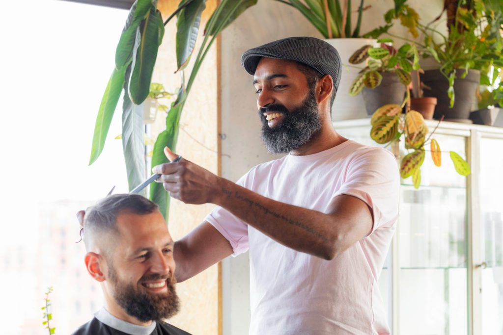 A smiling male barber with a dark beard and a grey flat cap uses scissors and a comb on a smiling customer's hair in a brightly lit salon filled with green indoor plants.