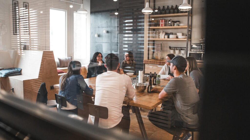 A candid view through a glass pane of a group of colleagues sitting around a long wooden table in a modern, open-plan office kitchen or dining space, talking and sharing drinks.