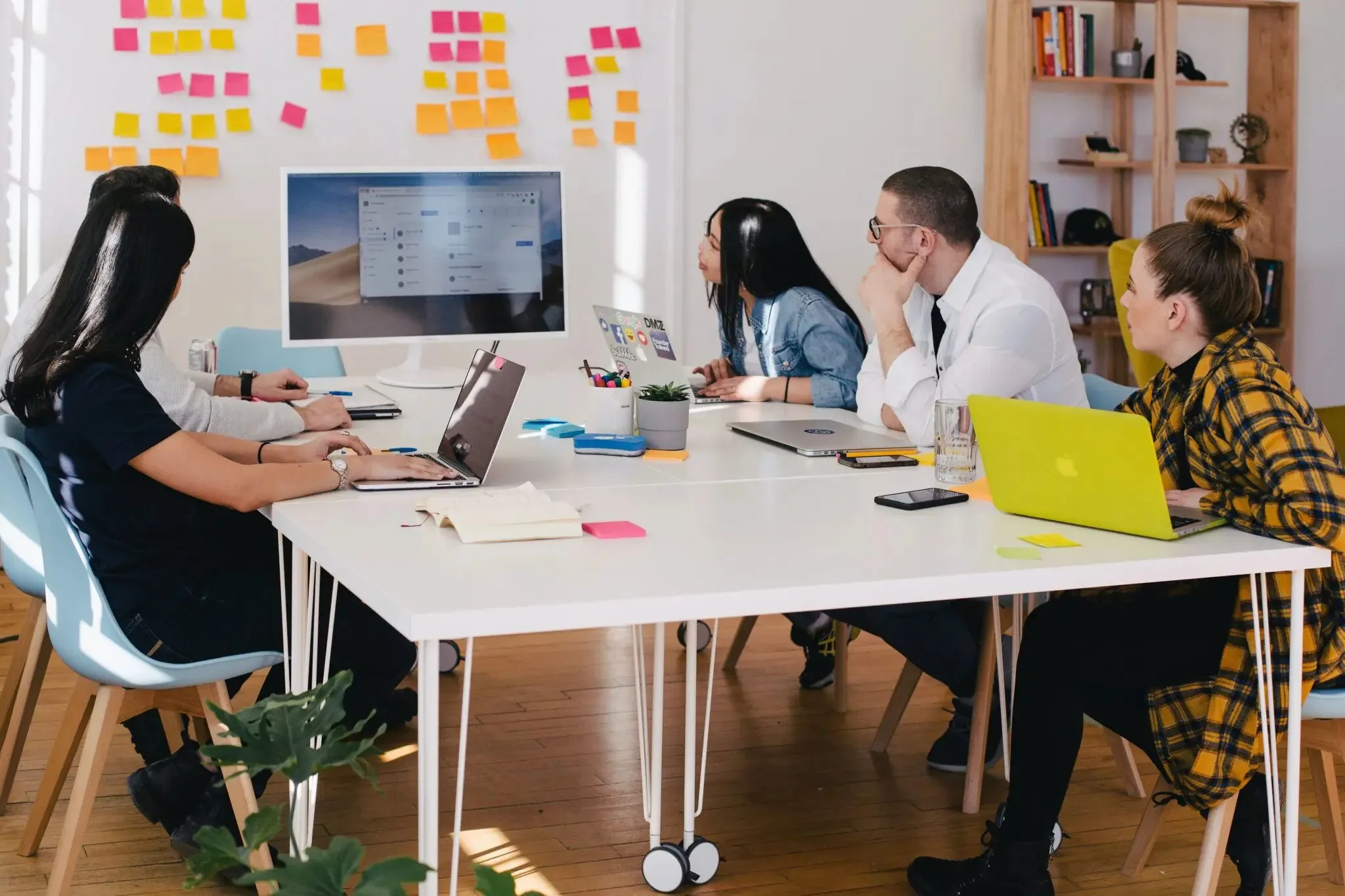A team of five planning around a white table with laptops, a wall of sticky notes behind them