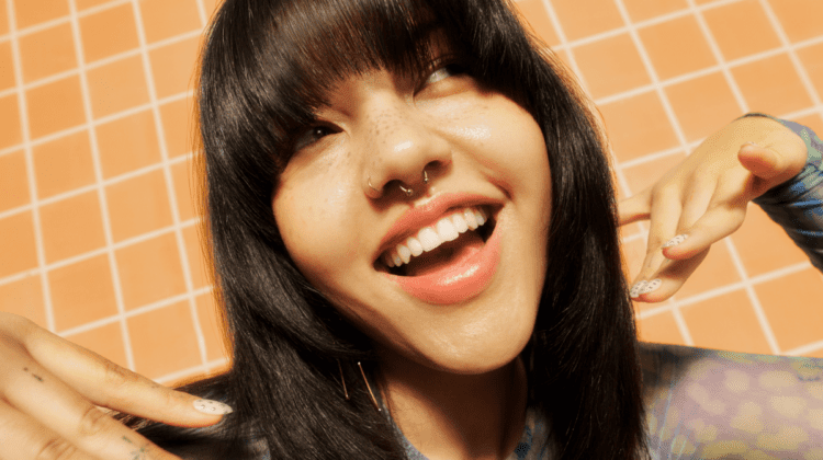 Smiling person with long dark hair, fringe, and nose ring against an orange tiled background.