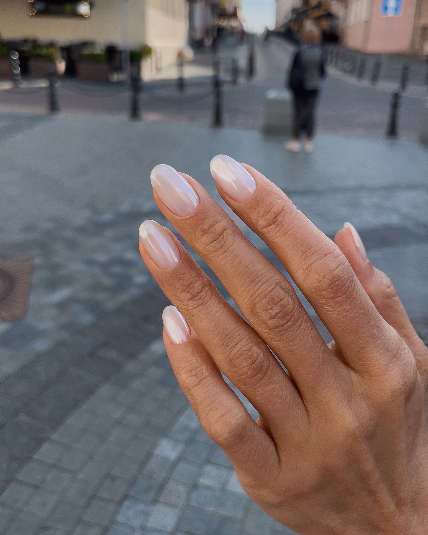 Hand with milky white, buffed nails held up against a blurred city street background. Credits @sansilt
