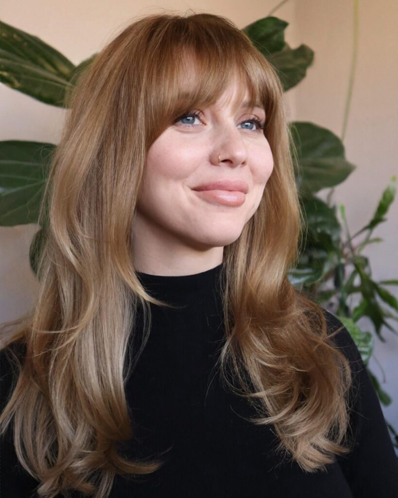 A woman with honey-blonde hair styled in eyebrow-grazing Birkin bangs, showcasing a tousled, effortless French-girl aesthetic.