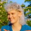 [ALT_TEXT: A young woman with a curly white blonde mullet haircut sitting outside on a sunny day.]
