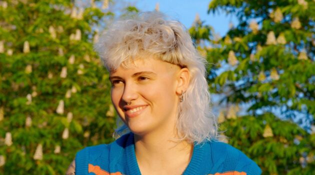 [ALT_TEXT: A young woman with a curly white blonde mullet haircut sitting outside on a sunny day.]