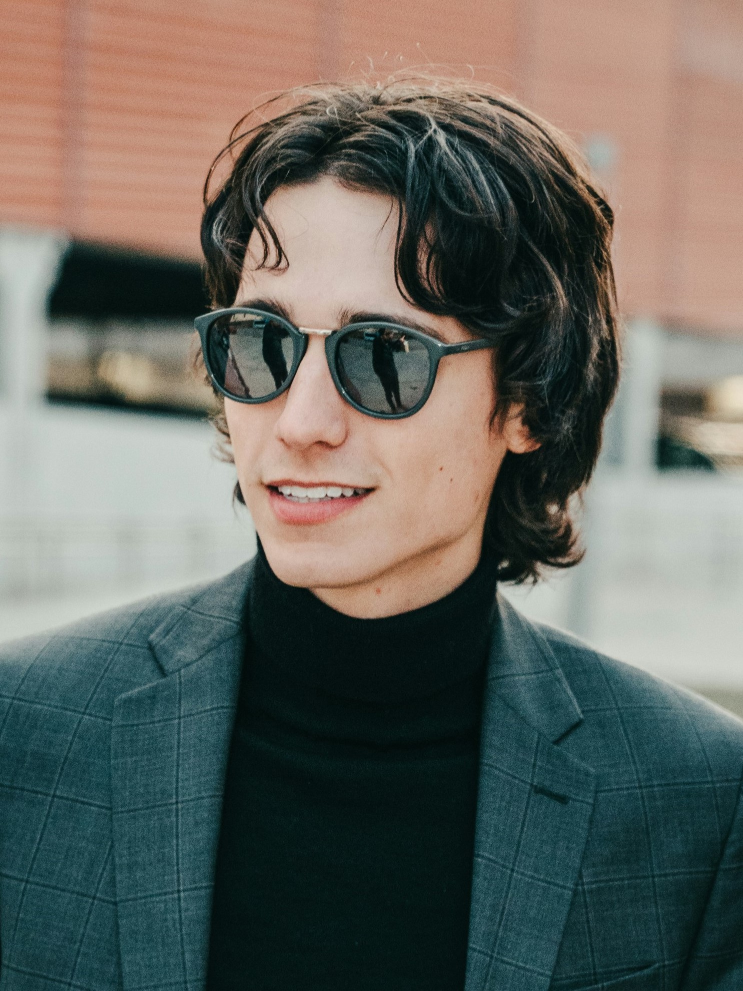 A young man with a retro mullet haircut wearing a grey suit jacket and sunglasses standing outside smiling.