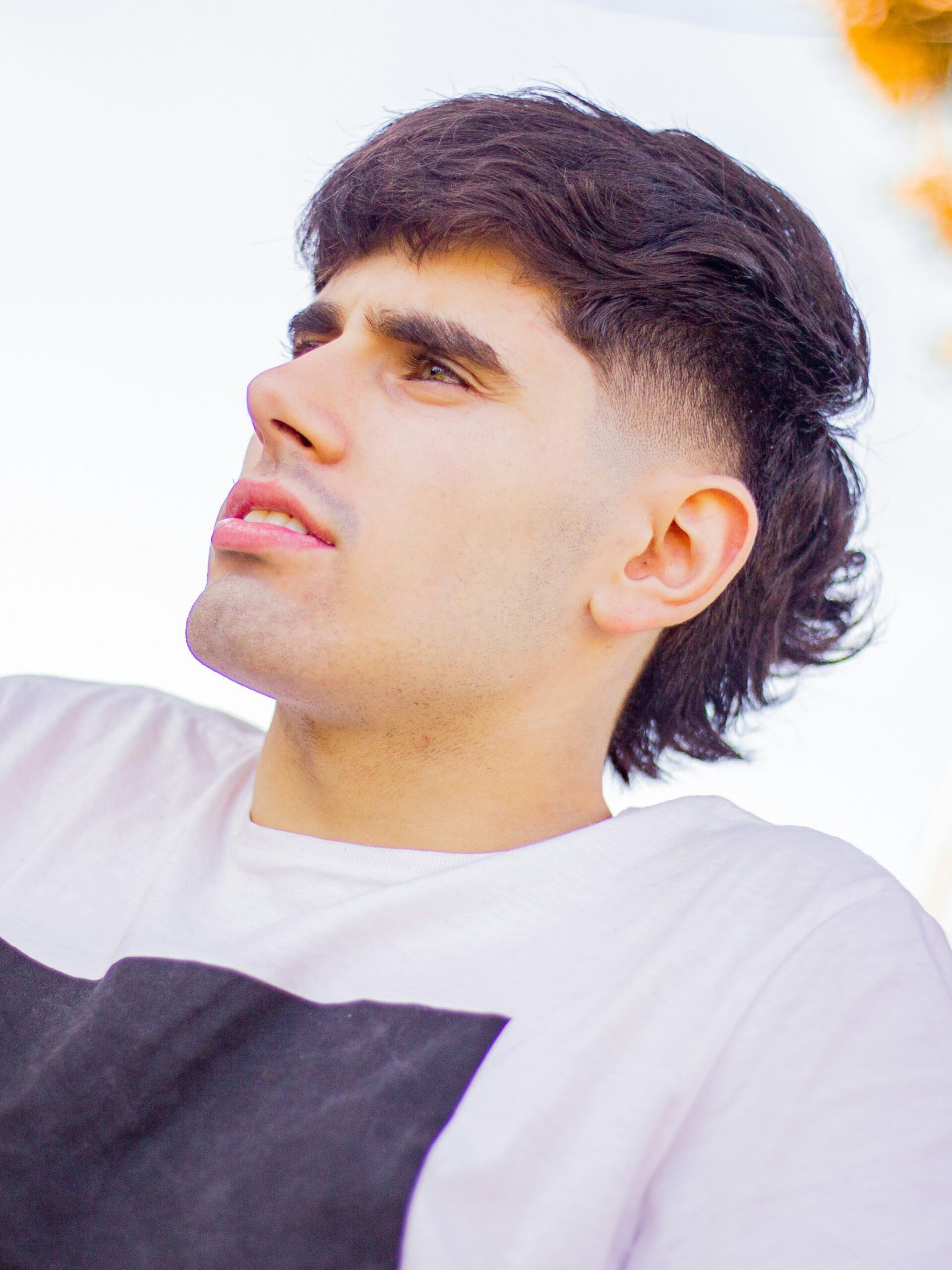 A young man with a sharp fade mullet haircut wearing a white t-shirt sitting by a road on a bright day.