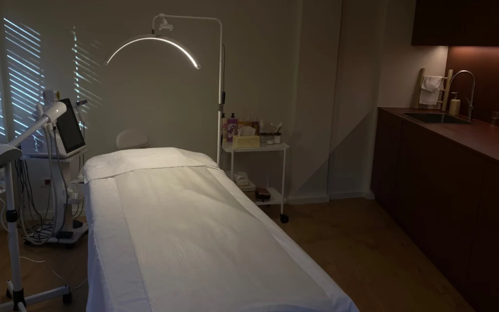 A focused treatment room with a white bed beneath an arc LED lamp, professional aesthetics equipment with a digital screen, skincare products on a trolley, and dark wood cabinetry with a sink along the far wall.