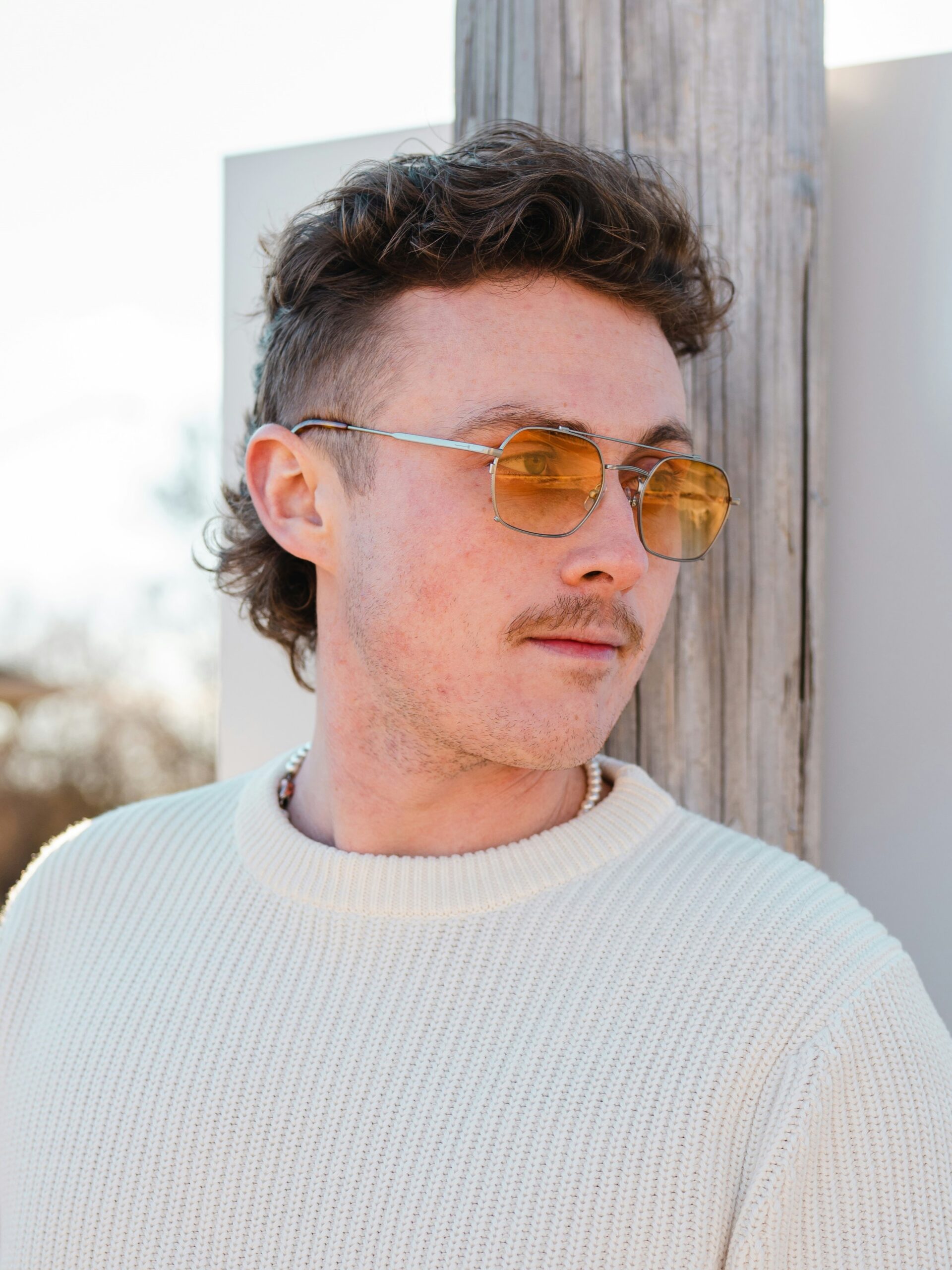 A young man with a curly mullet haircut leaning on a wall outside on a sunny day.