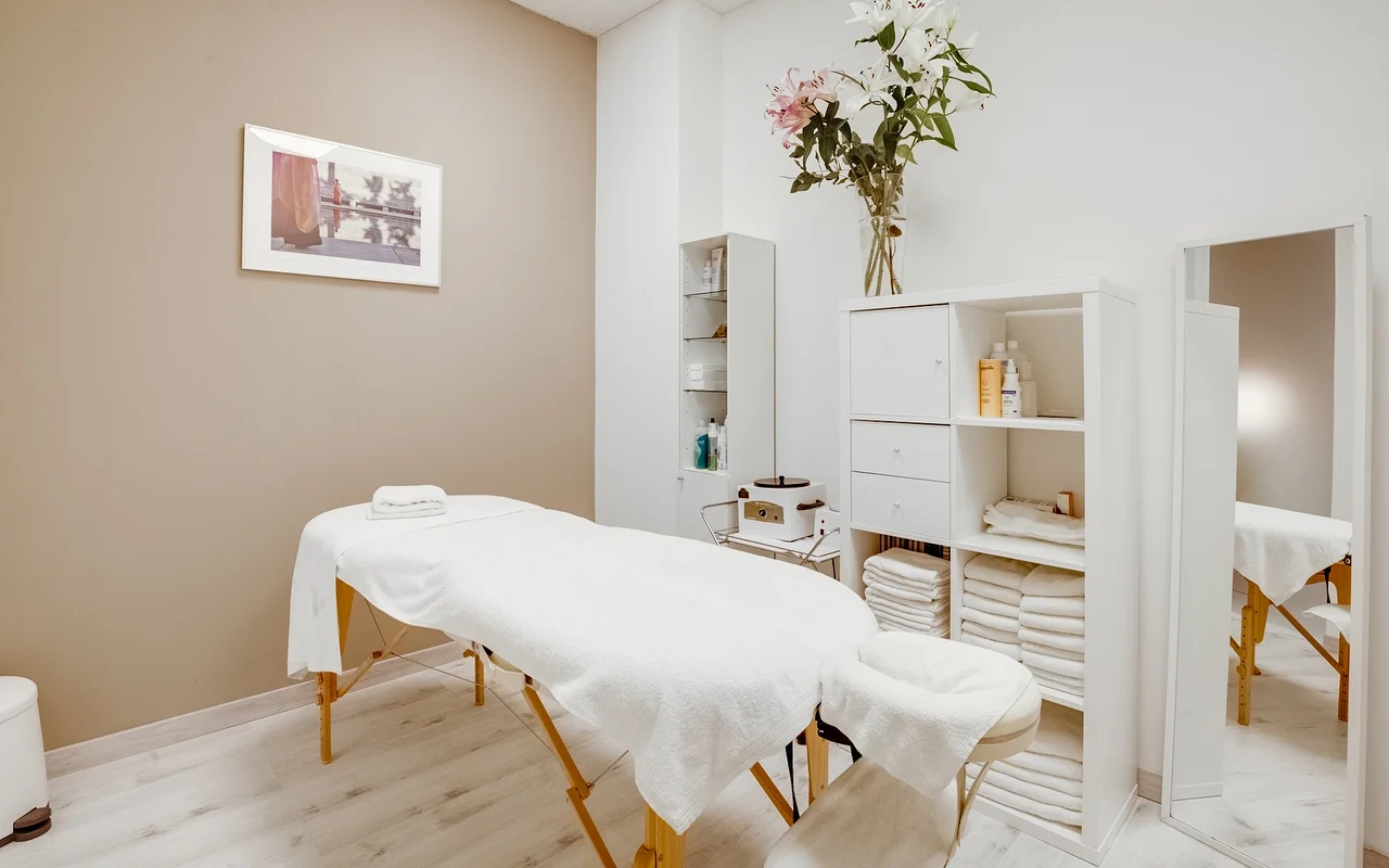 A serene treatment room with a white massage table draped in fresh towels. The room has neutral beige and white walls, a white storage cabinet stocked with folded towels, and a tall glass vase with white and pink lilies.