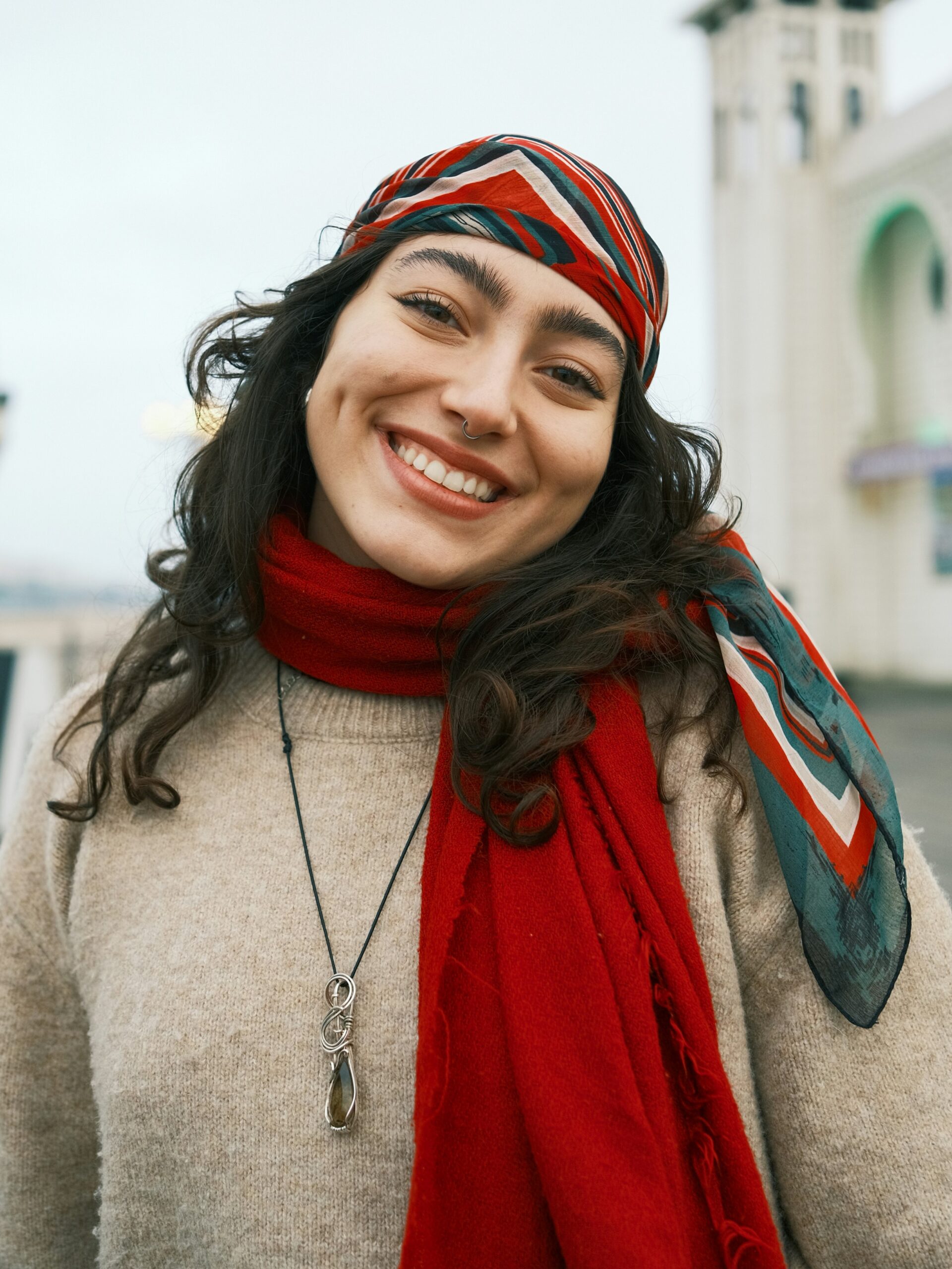 Smiling woman standing outside on a pier with long dark hair wearing a striped red, white and green scarf on her head and a red scarf around her neck