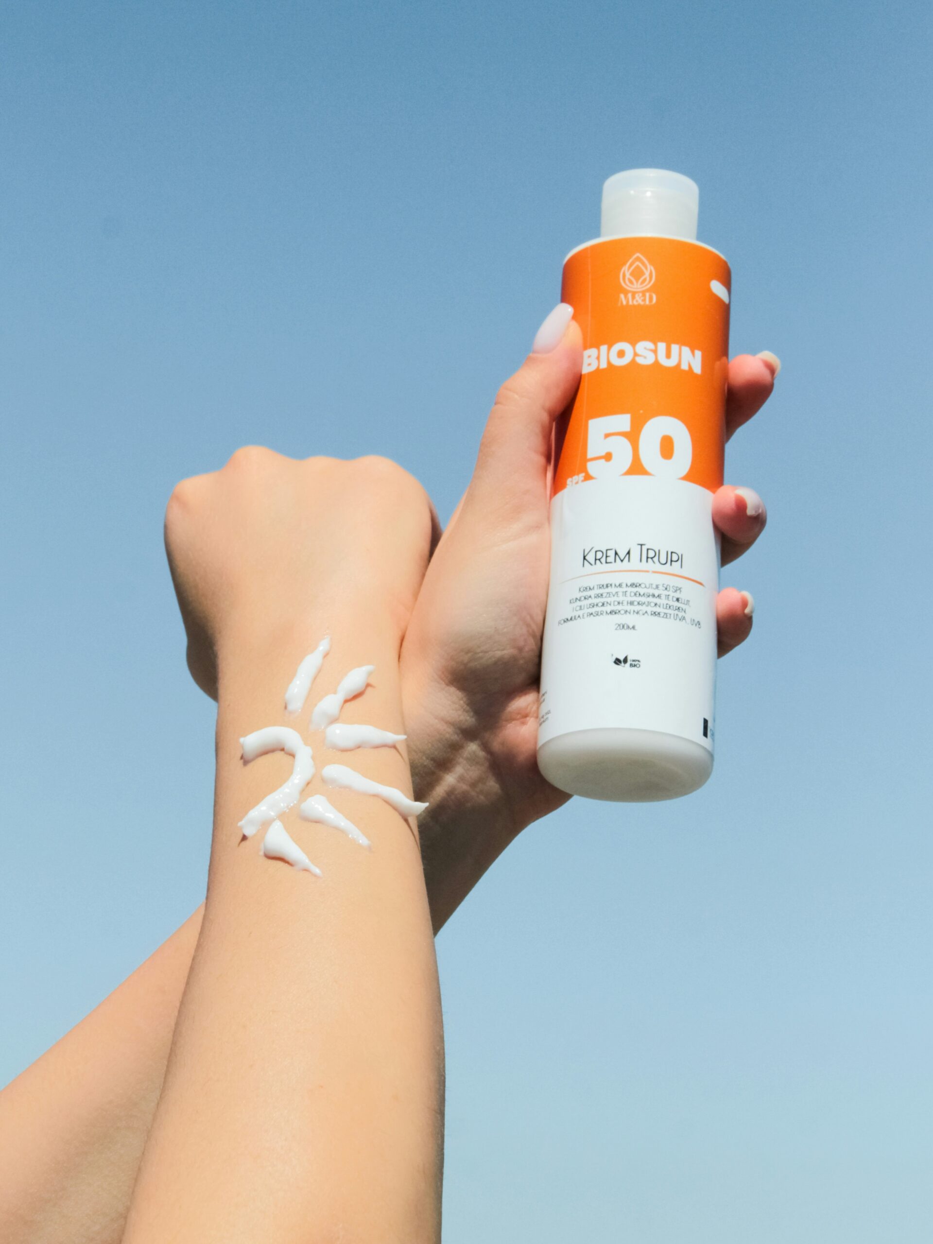 Close-up of a woman’s arms against a blue sky, one hand holds a bottle of suncream and one forearm has suncream in the shape of a sun