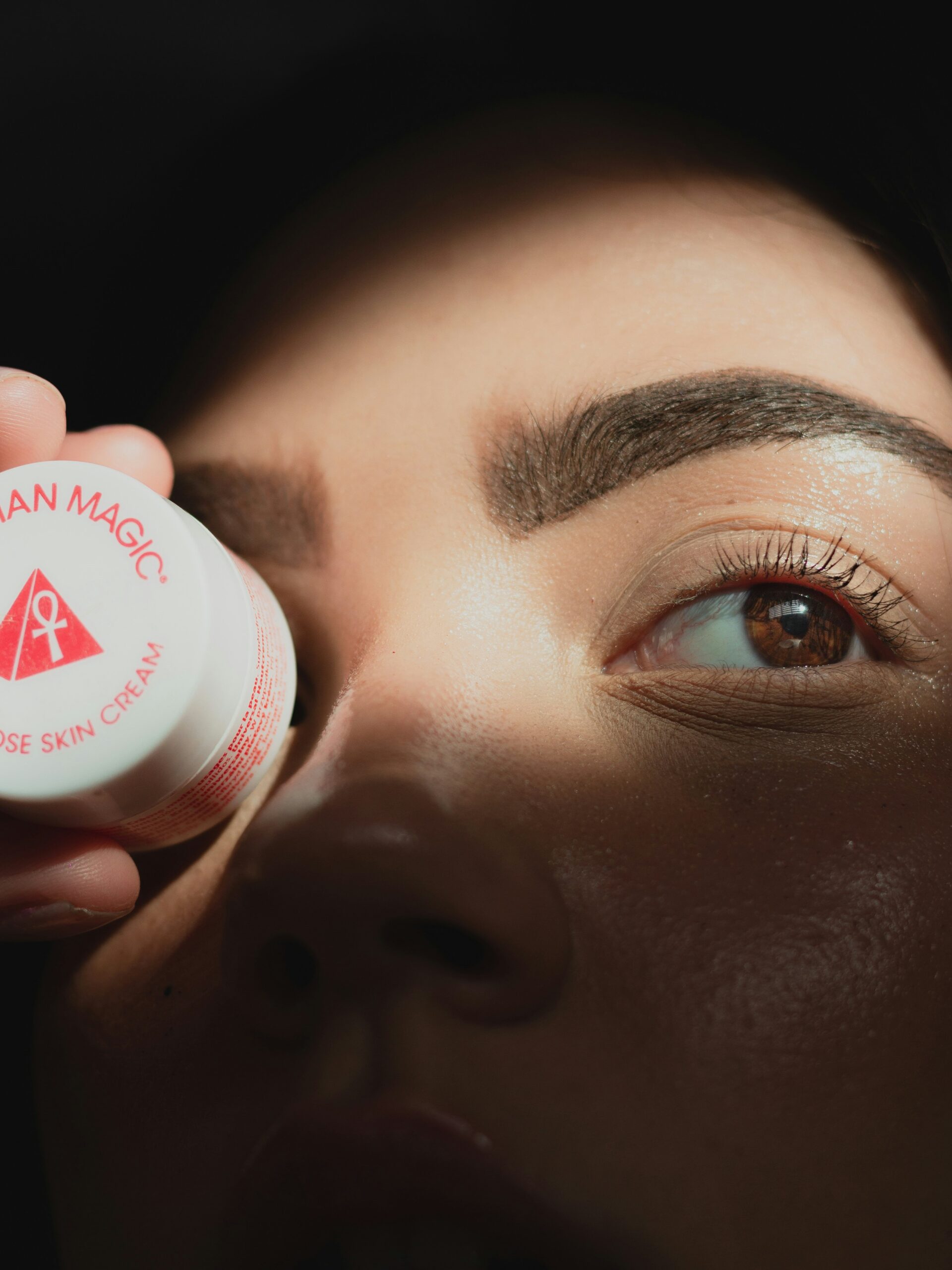 A close-up of a woman’s face with microblading on her brows holding a small round pot of cream in front of one eye