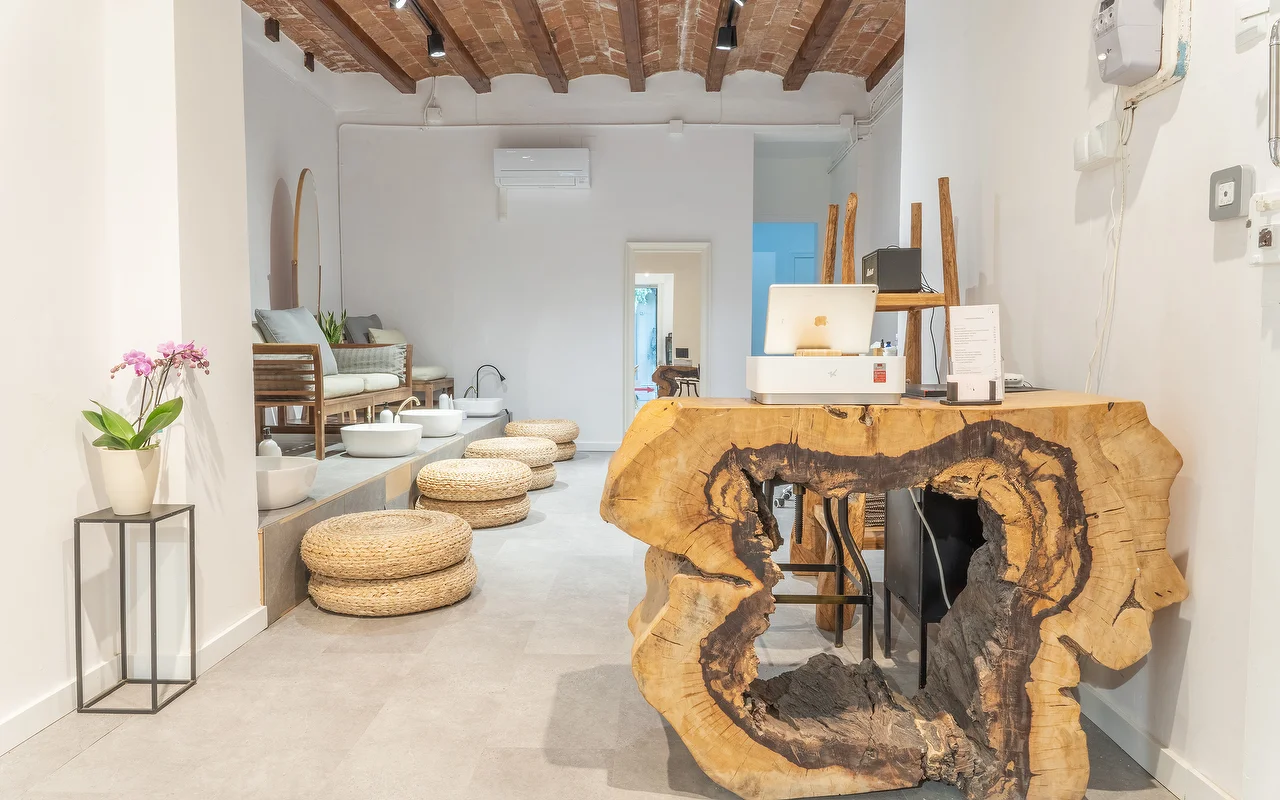 A minimalist wellness space featuring a reception desk made from a large, raw cross-section of a tree trunk. In the background, there is a pedicure station with woven floor cushions and white basins under a rustic beamed ceiling.