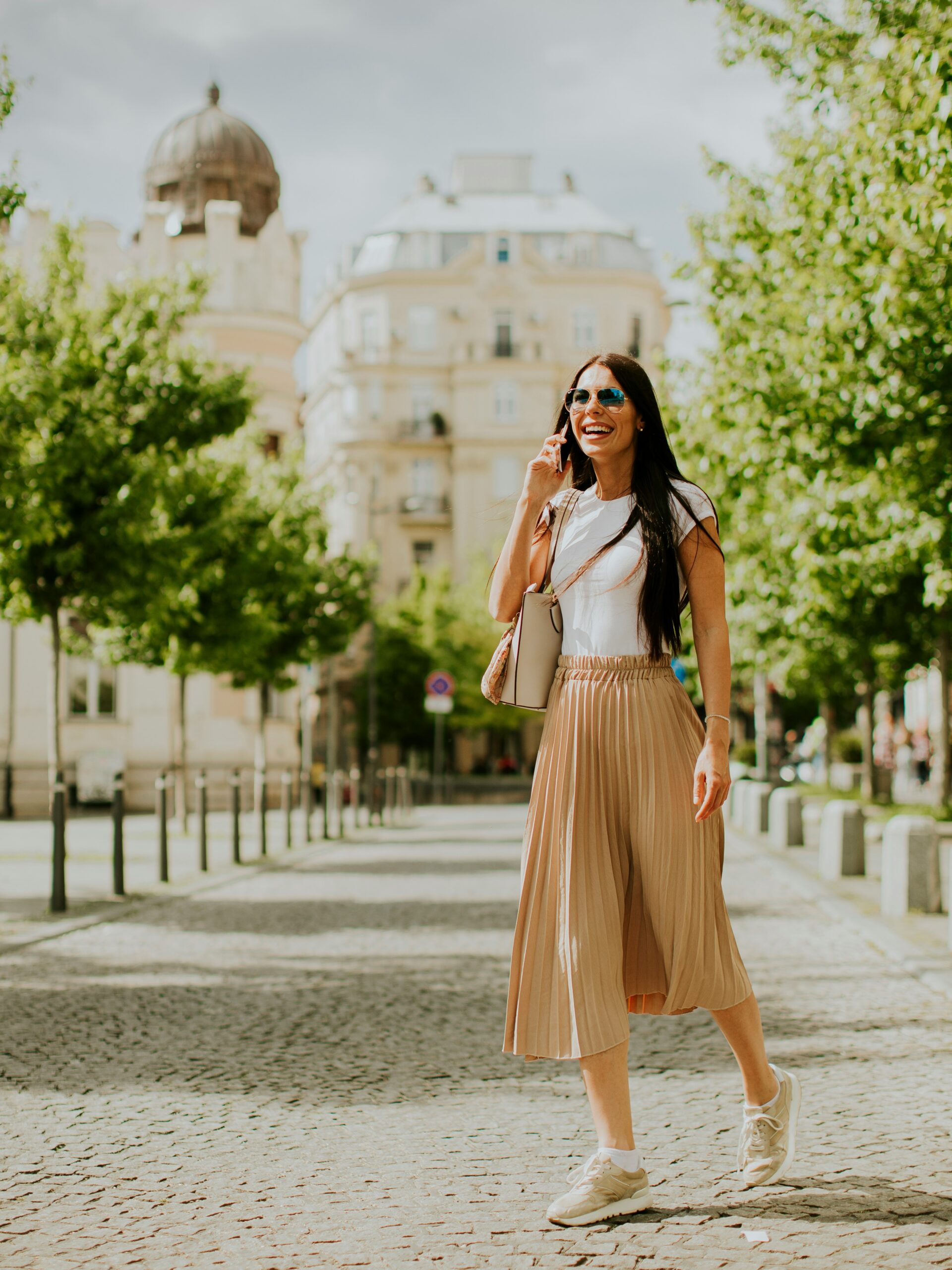 A smiling woman chats on her phone walking down a cobbled street wearing a loose white t-shirt and flowing mid-length skirt