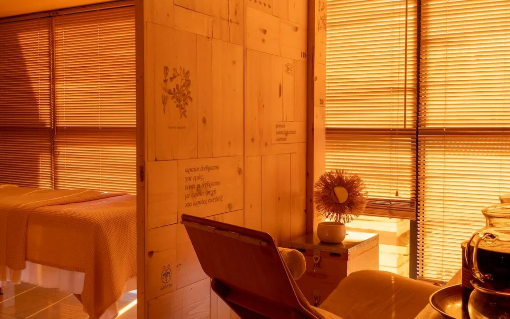 A warm, amber-lit spa treatment room with natural wood panelling, botanical illustrations and Greek text etched into the wall, a treatment bed draped in a waffle-weave blanket, and the APIVITA logo in the corner.
