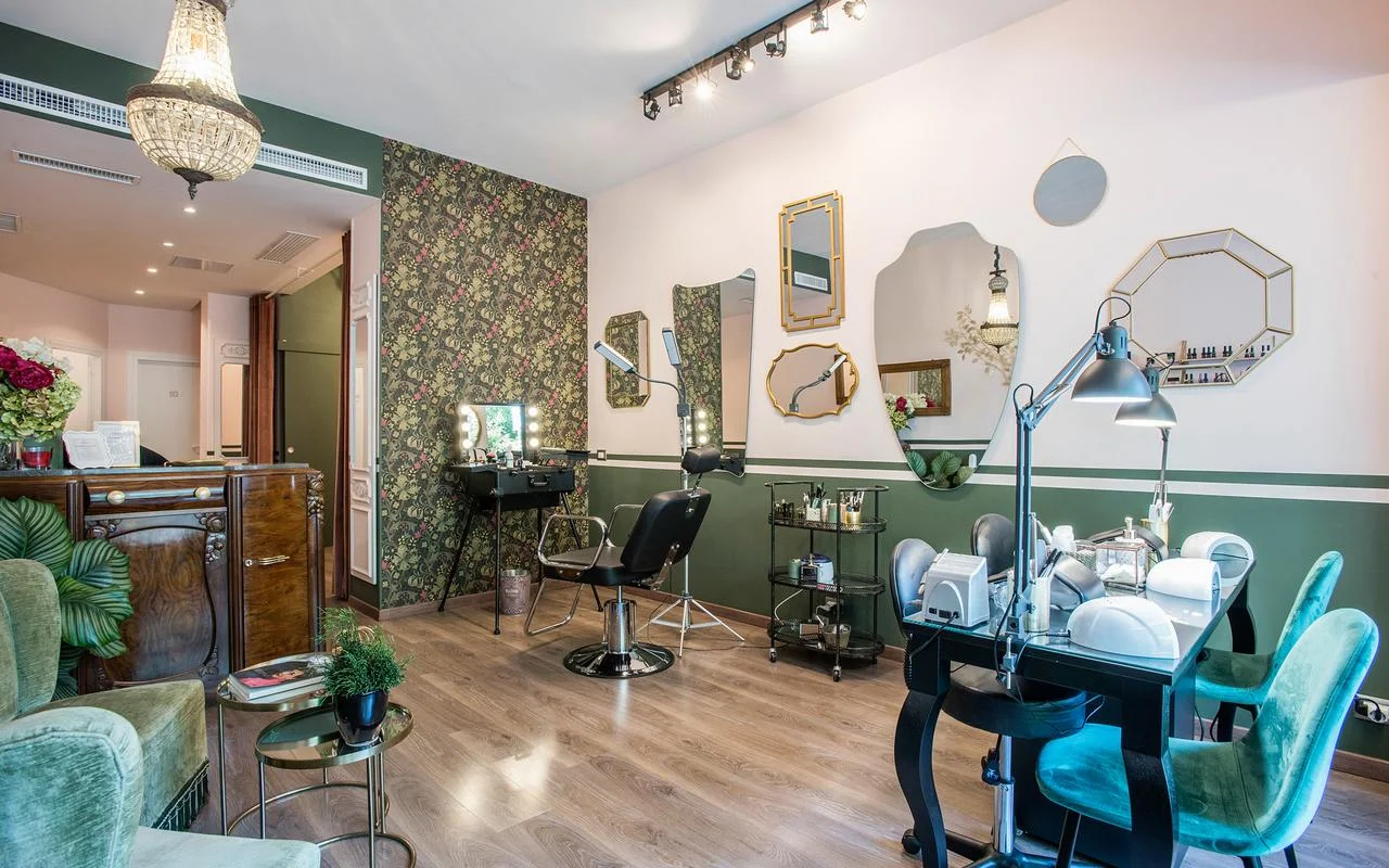 A vintage-chic salon with white and forest green walls. It features floral patterned wallpaper, an assortment of irregularly shaped gold-framed mirrors, a professional make-up station, and plush green velvet seating.