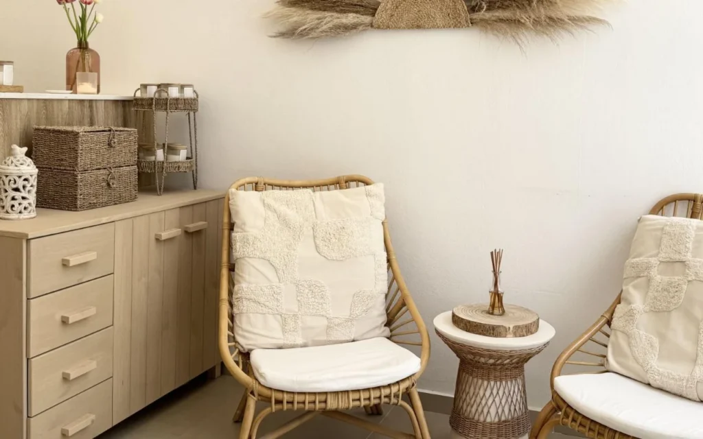 A calming waiting area with rattan armchairs, textured cream cushions, a wicker side table with a reed diffuser, woven storage baskets, and dried pampas grass on the wall above pale wood cabinetry.