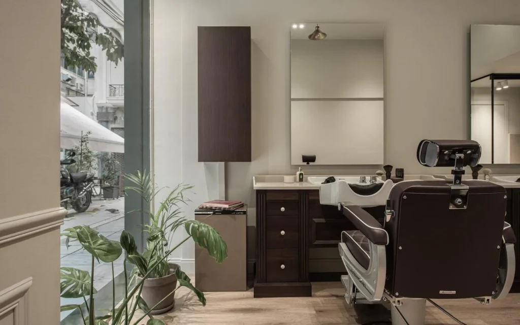 A refined barbershop with a dark brown leather barber chair, walnut-wood cabinetry, a large window looking out onto a Kolonaki street, and a monstera plant adding greenery to the minimalist space.