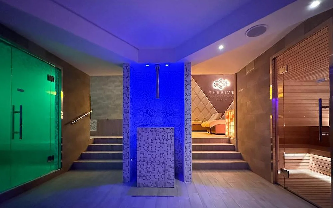 A luxury spa’s thermal area with dramatic blue and green atmospheric lighting. The space features a wooden Finnish sauna, a steam room entrance, and a central mosaic-tiled feature with steps leading to relaxation zones.