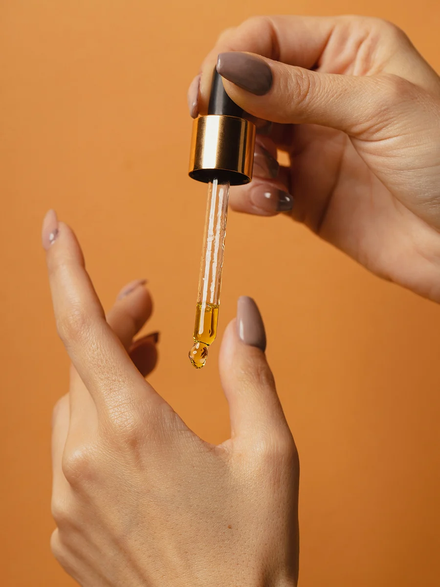 A close-up of two manicured hands with one hand holding a glass dropper with cuticle oil dripping out