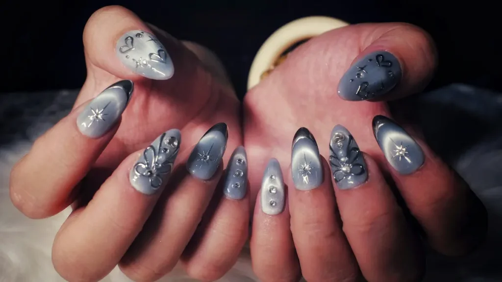 Close-up of a woman's hands showing intricate acrylic nails with a grey cats-eye design, silver hearts and stars, and sparkling gemstone embellishments