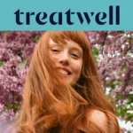 A woman with long copper-red hair and a fringe smiling in the sunshine, with pink blossom trees in the background. The Treatwell logo sits on a teal banner above.
