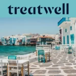 The Little Venice waterfront in Mykonos Town: whitewashed houses with colourful wooden balconies at the sea's edge, the turquoise Aegean beyond, and white and teal café tables set out across a stone-paved terrace in the foreground.
