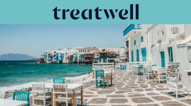 The Little Venice waterfront in Mykonos Town: whitewashed houses with colourful wooden balconies at the sea's edge, the turquoise Aegean beyond, and white and teal café tables set out across a stone-paved terrace in the foreground.