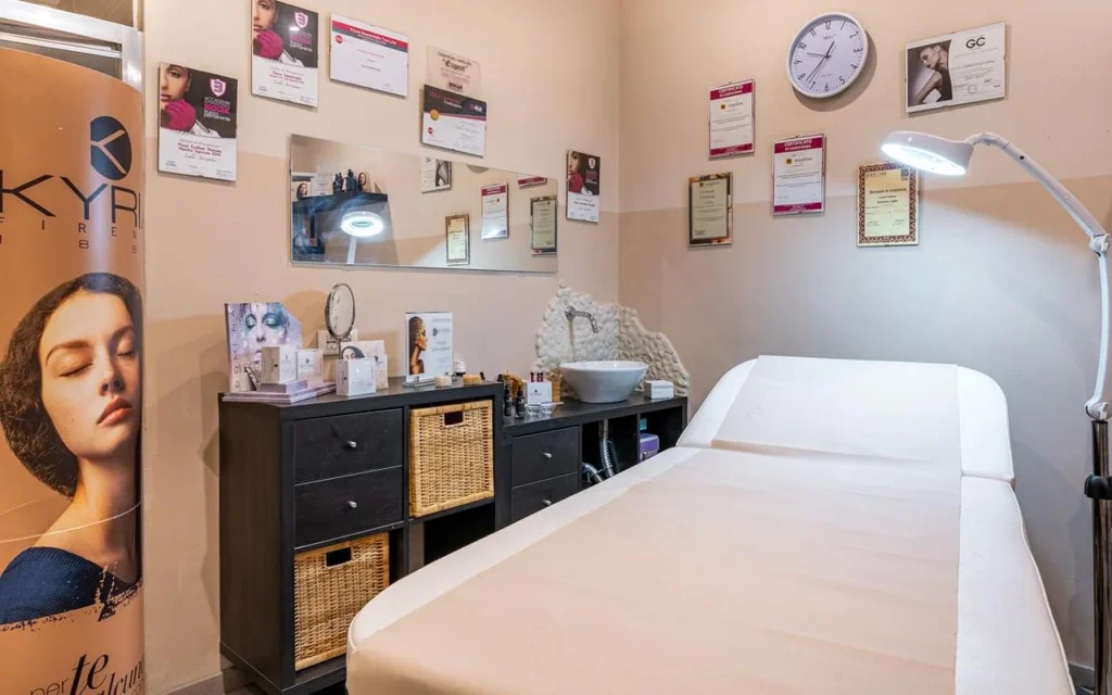 A treatment room at Oltre la bellezza with a white treatment bed, adjustable magnifying lamp, and walls lined with professional certificates and diplomas. Dark wicker storage units sit beneath a mirror, with skincare products arranged on the counter.