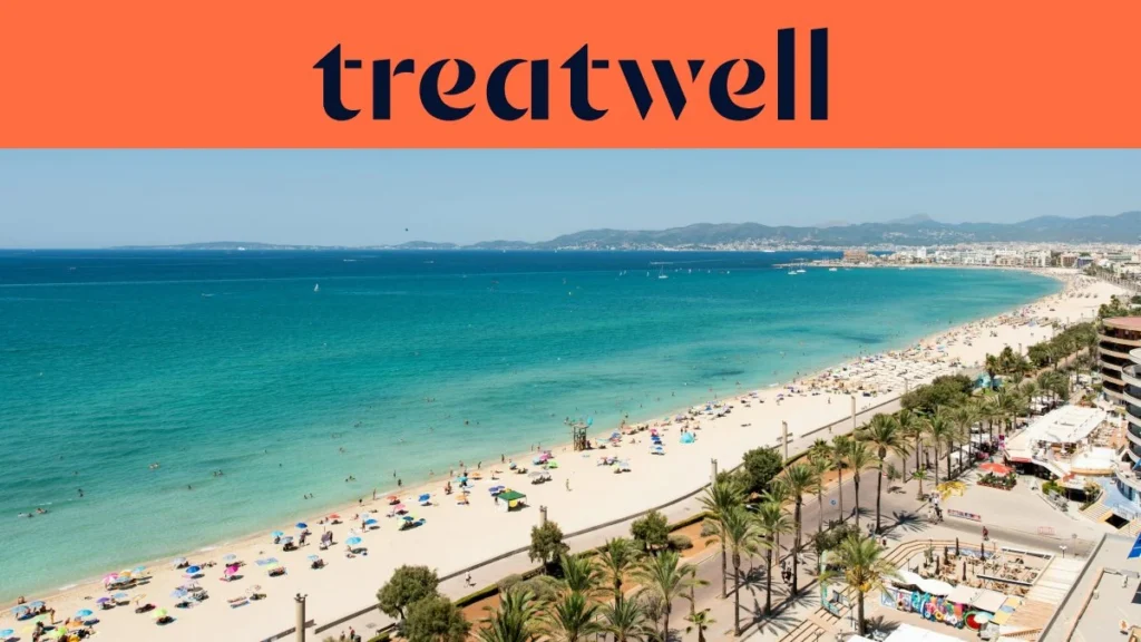 An aerial view of Playa de Palma in Mallorca, with a long curve of golden sand, rows of beach umbrellas, calm turquoise water, and a palm-lined promenade running alongside the city, with hills in the distance under a clear blue sky.