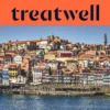 The Ribeira waterfront in Porto, with rows of colourful houses and terracotta rooftops rising up the hillside from the Douro river and the Clérigos tower on the skyline under a clear blue sky.