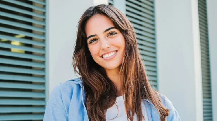 A woman with long dark hair and well-defined brows stands outside smiling in front of a building with green shutters