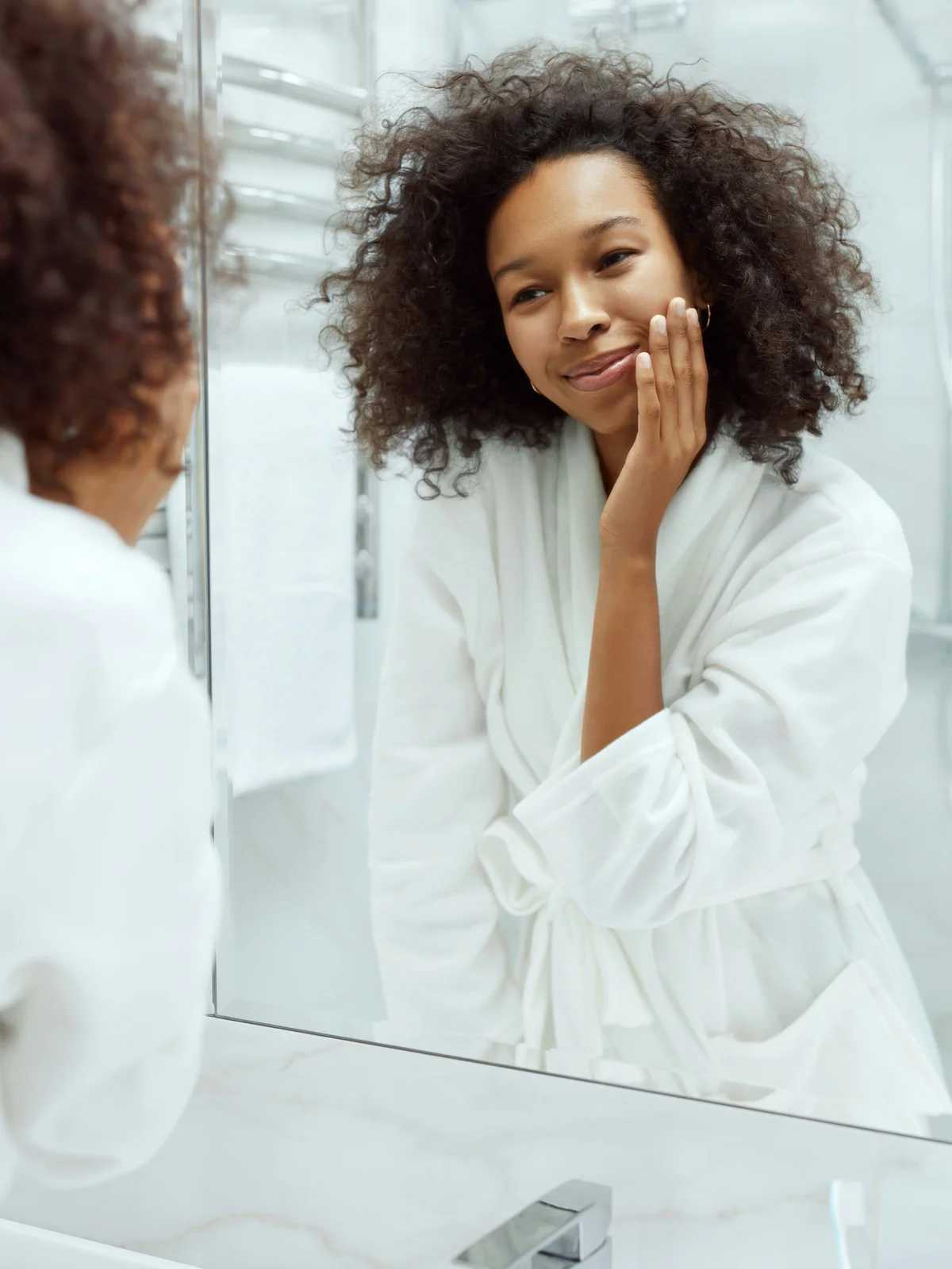 A woman wearing a white bathrobe looks at herself in the mirror and lightly strokes her face showing her smooth, clear skin