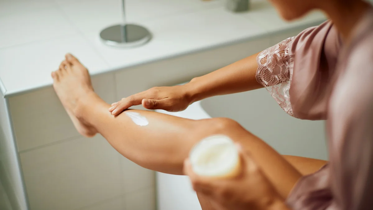 Over-the-shoulder shot of a woman wearing a lilac silk robe applying moisturiser to her leg as she rests it on the counter in her bathroom