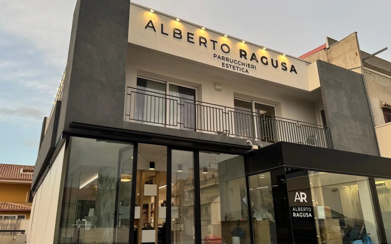 The exterior of Alberto Ragusa at dusk, a two-storey salon with a large illuminated sign reading 'Alberto Ragusa Parrucchieri Estetica' above floor-to-ceiling glass windows, warm light glowing from inside.