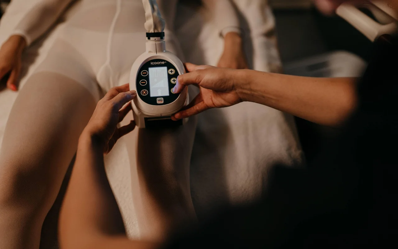 A body-contouring treatment at Beauty Room Romina Bongiovanni, with a therapist holding an icoone device against the leg of a client in a white treatment bodysuit, shown in warm low light.