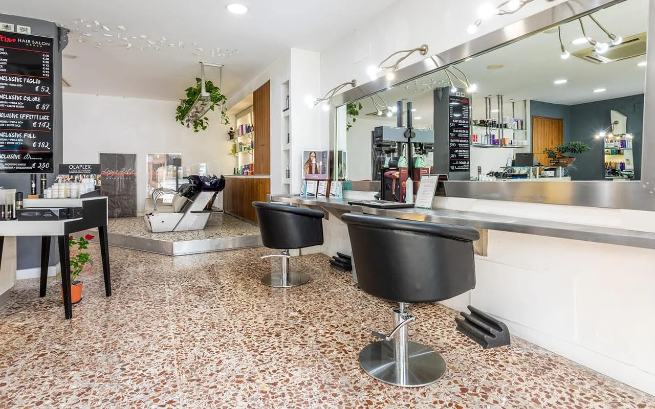The interior of Hair Salon Luigi Zito, with a terrazzo floor, two black styling chairs facing a long mirror, chalkboard price lists on the wall, a product display and a backwash basin in the background.