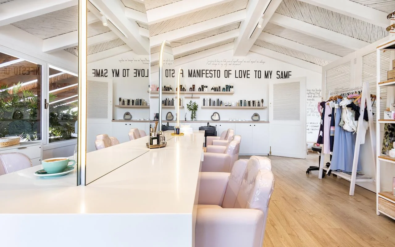 The bright interior of Milk & Roses at Puente Romano, Marbella, with a white styling counter, pale pink chairs, a beamed ceiling and product shelves along the wall.