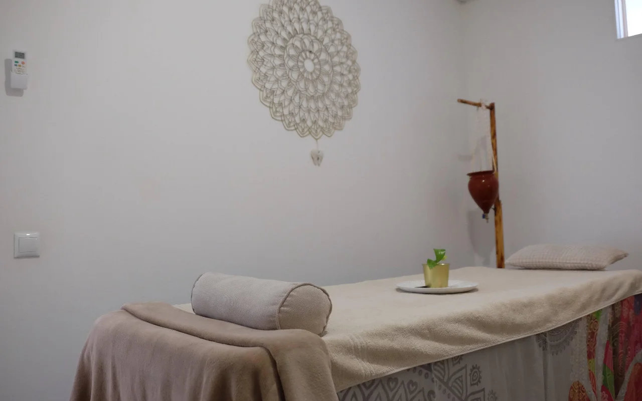 A calm massage room in soft neutral tones, with a towel-draped massage bed and bolster, a large crochet mandala hanging on the wall, and a wooden stand holding a suspended terracotta pot.