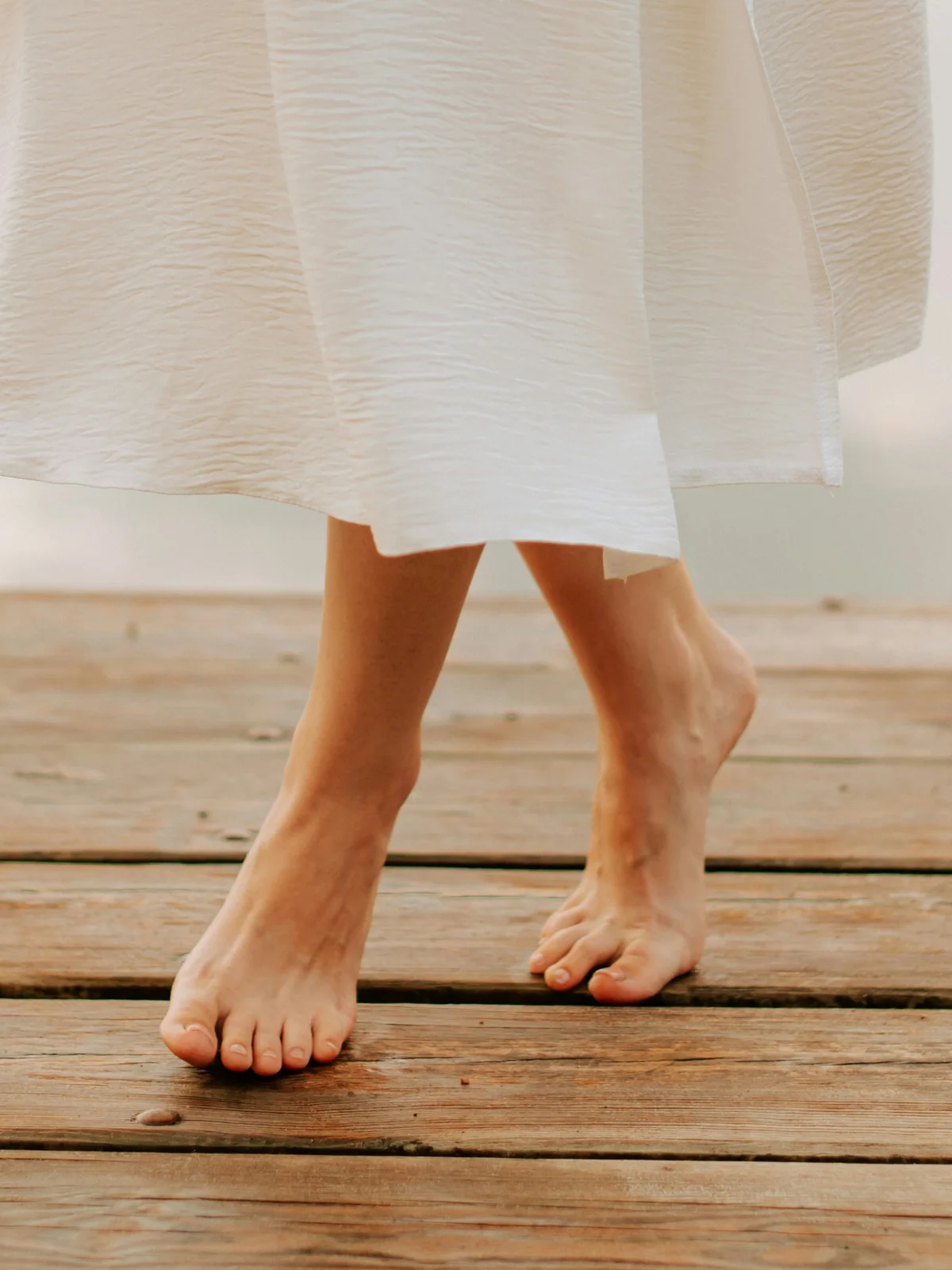 Close-up of a woman’s lower legs with healthy bare feet and a flowing white skirt as she takes a step forward on a wooden deck by a lake
