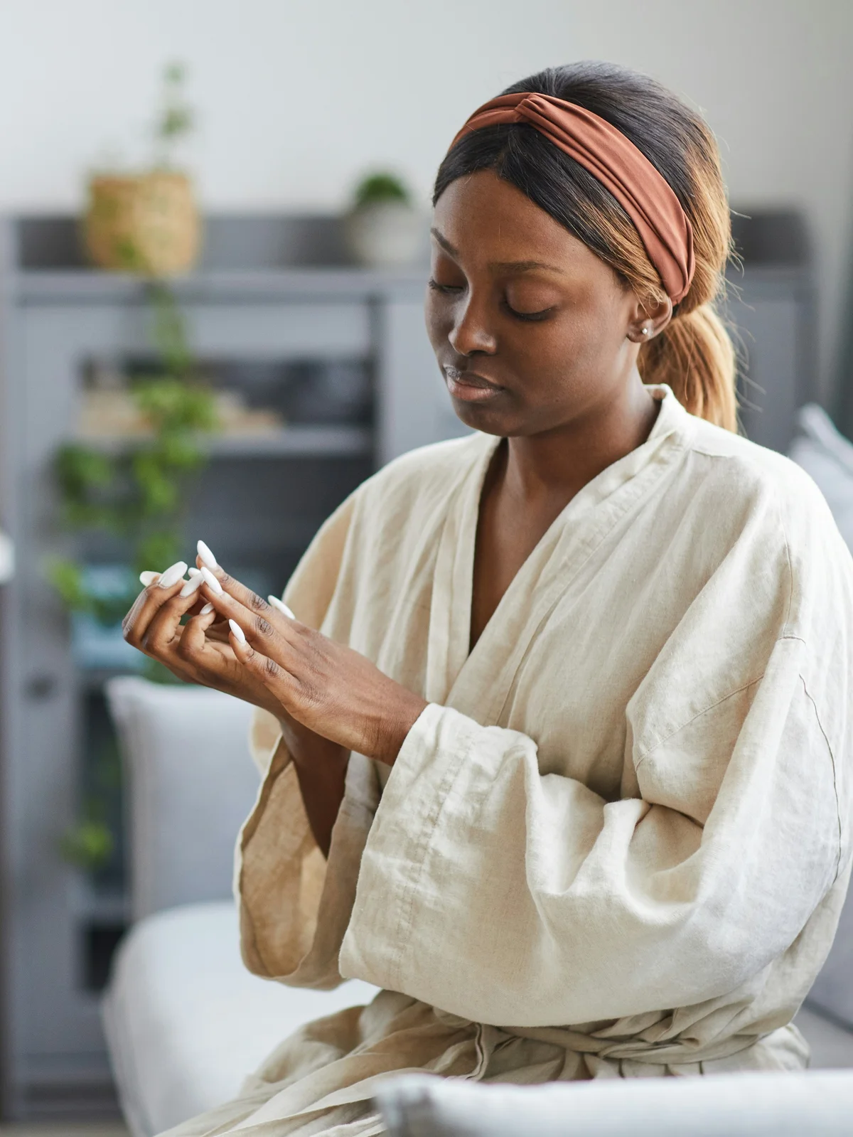 A woman sits at home wearing a light robe and headband, she carefully massages cuticle oil into her manicured fingers