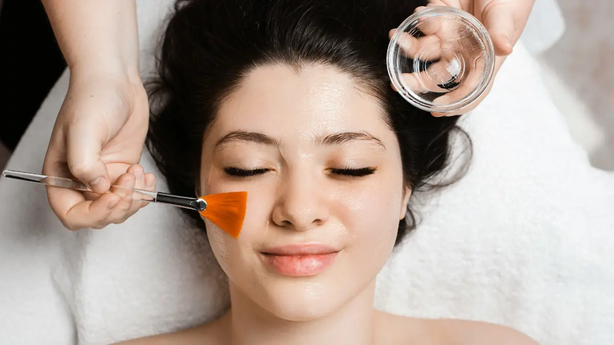 Top view of a client’s face with eyes closed as a therapist’s hands apply a soothing face mask using a brush