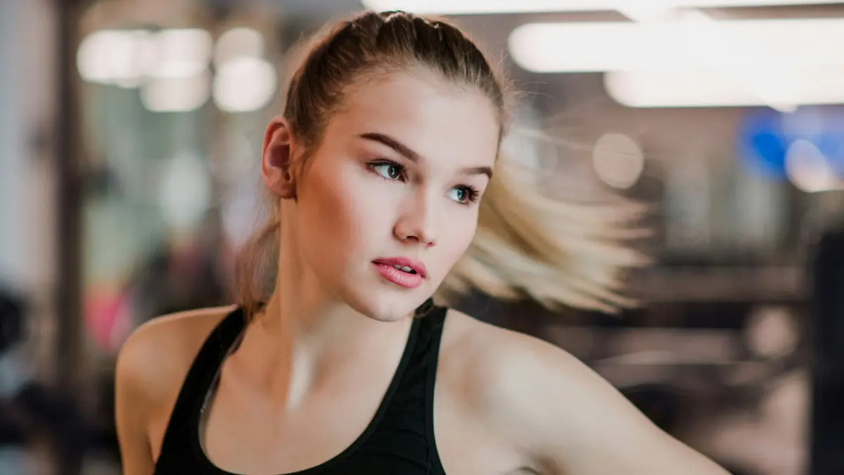 A young woman wearing a gym vest top moves sideways as her ponytail flies out to the side