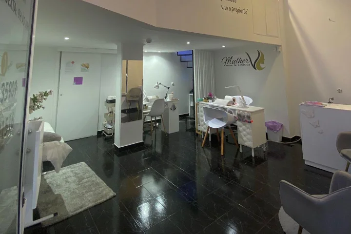 The interior of a beauty centre with a polished dark tiled floor, a white manicure station and softly lit treatment areas, the salon's Mulher logo on the back wall and a facial bed visible behind a glass partition.