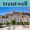 A sunny view over Ibiza Town, with whitewashed buildings climbing to the cathedral and old fortress walls of Dalt Vila on the hill. A tree-lined harbour promenade runs across the foreground beneath a clear blue sky.