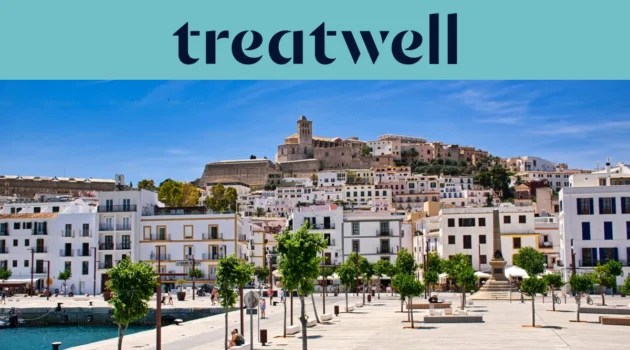 A sunny view over Ibiza Town, with whitewashed buildings climbing to the cathedral and old fortress walls of Dalt Vila on the hill. A tree-lined harbour promenade runs across the foreground beneath a clear blue sky.