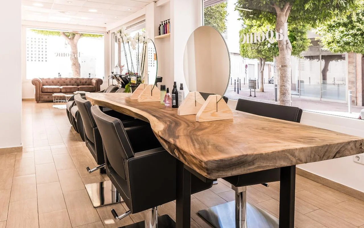 A bright hair salon built around a long live-edge wooden counter lined with black styling chairs. A round mirror and wooden product stands sit on the counter, a brown leather Chesterfield sofa waits at the back, and tall windows look out onto a leafy street.