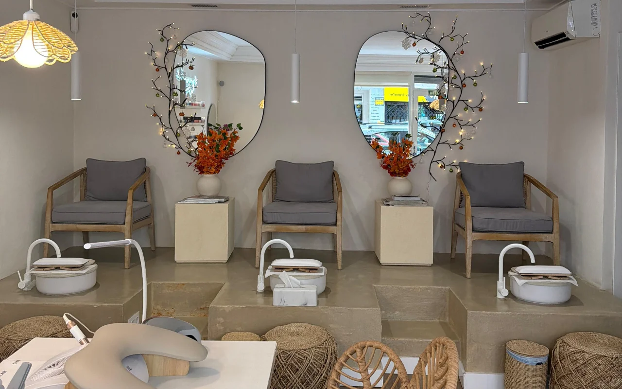 A bright, modern nail salon with two pedicure chairs on a raised concrete platform, each with a white foot bath and a grey cushioned armchair. Two organic-shaped mirrors dressed with fairy-lit branches and orange blooms hang above, with a manicure station in the foreground.