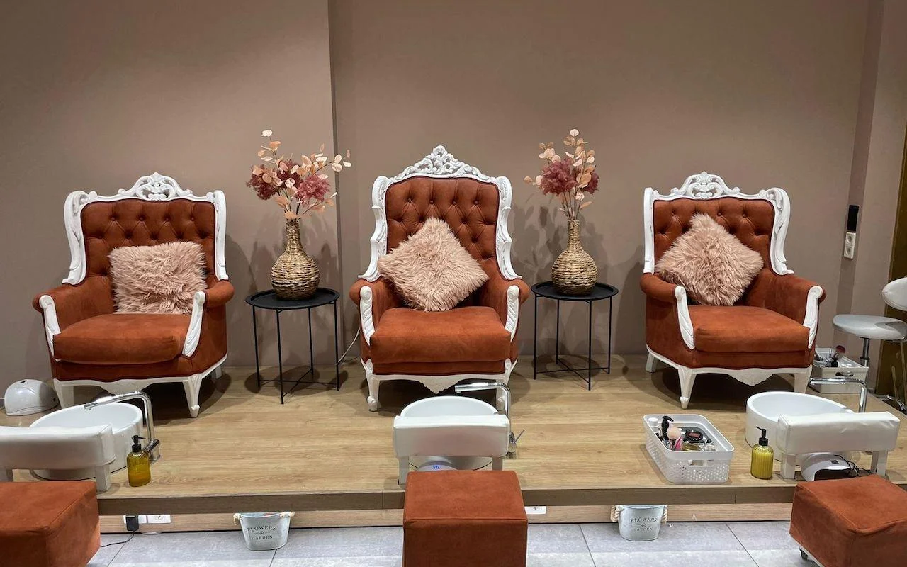 Three ornate white-framed armchairs in burnt-orange velvet, raised on a wooden platform above white pedicure basins, with dried flowers and soft pink cushions set between the seats.